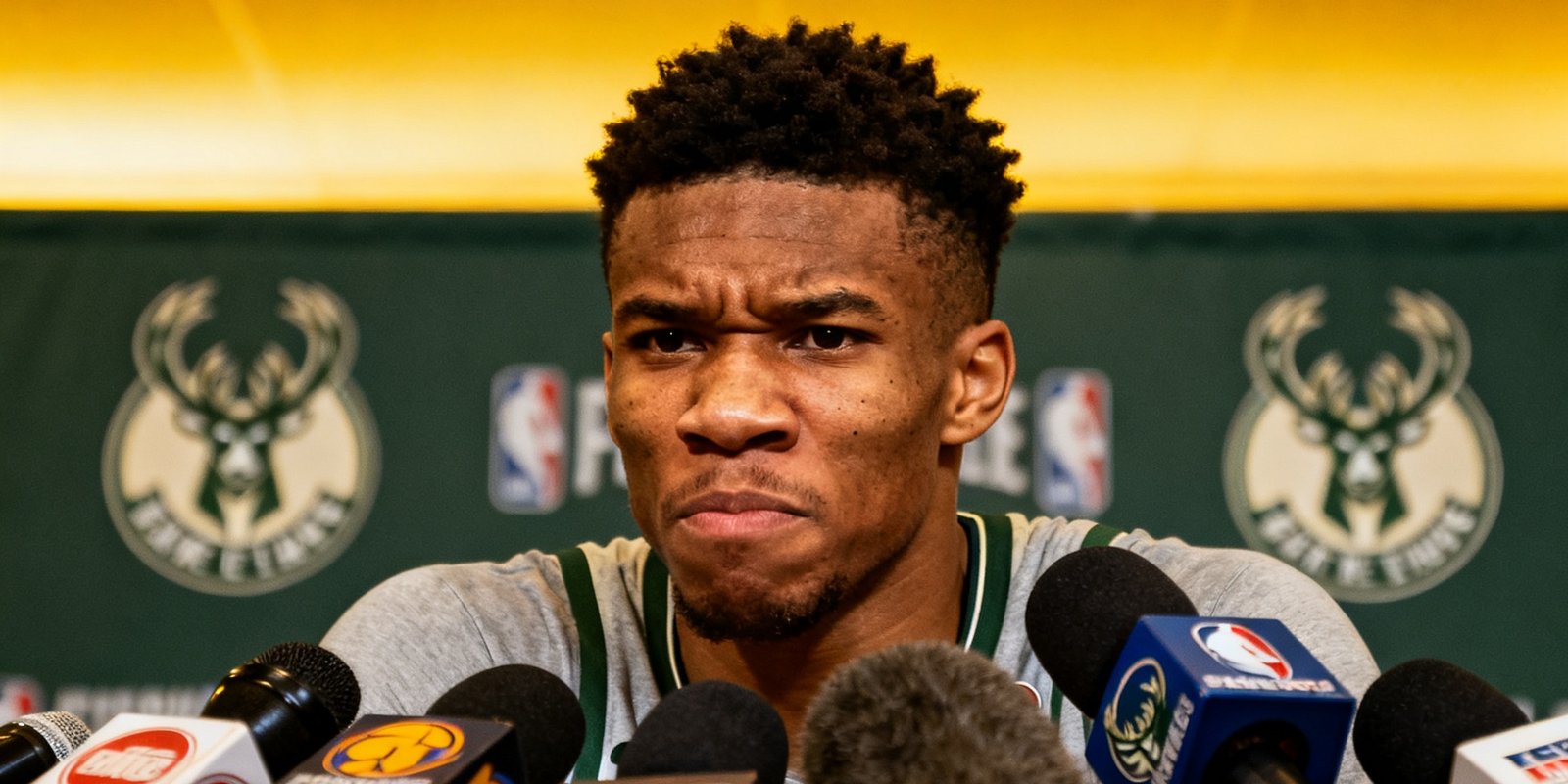 NBA转会市场新闻配图，字母哥扬尼斯·阿德托昆博在新闻发布会上面对众多记者话筒，表情严肃，背景是球队标志墙。